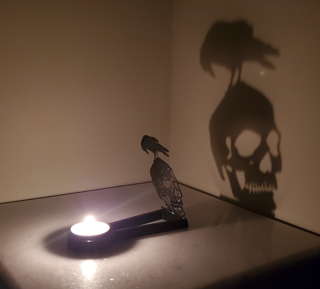 The Kealite Nevermore Shadow Caster set up with a tea light lit and a shadow of a raven perched on top of a skull is projected on the wall. 