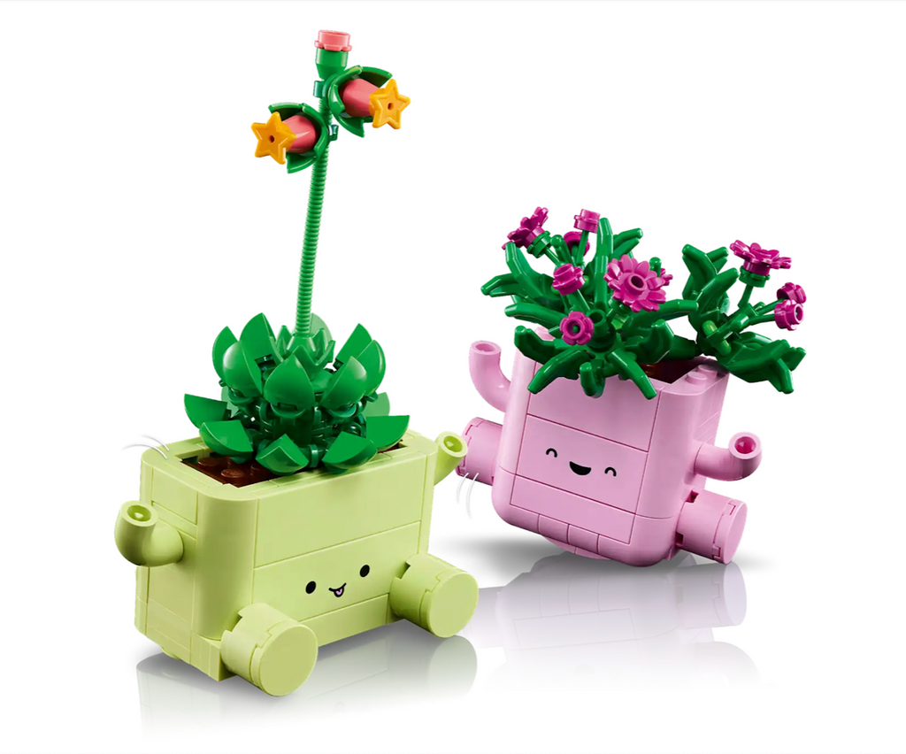 Two LEGO cute planters, one is light green with a green succulent and a tall flower stem with yellow and pink blossoms and the other is pink planter with dark green foliage with fuchsia flowers. 