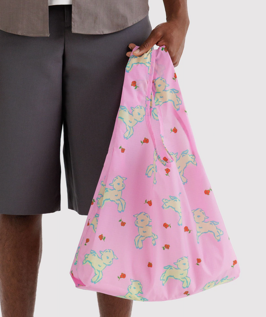 A person holding a full standard Baggu bag in the lambs print with cute little lambs on a pink background. 
