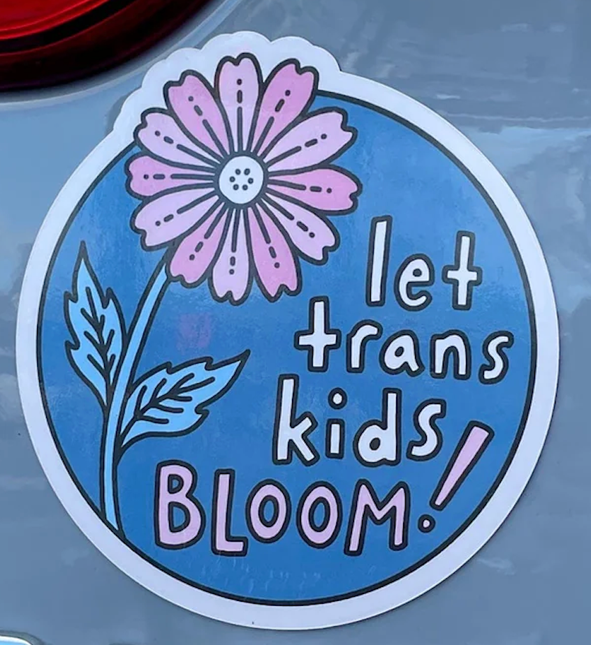 Round magnet with blue background and white lettering with a purple flower that reads "Let Trans Kids Bloom!"