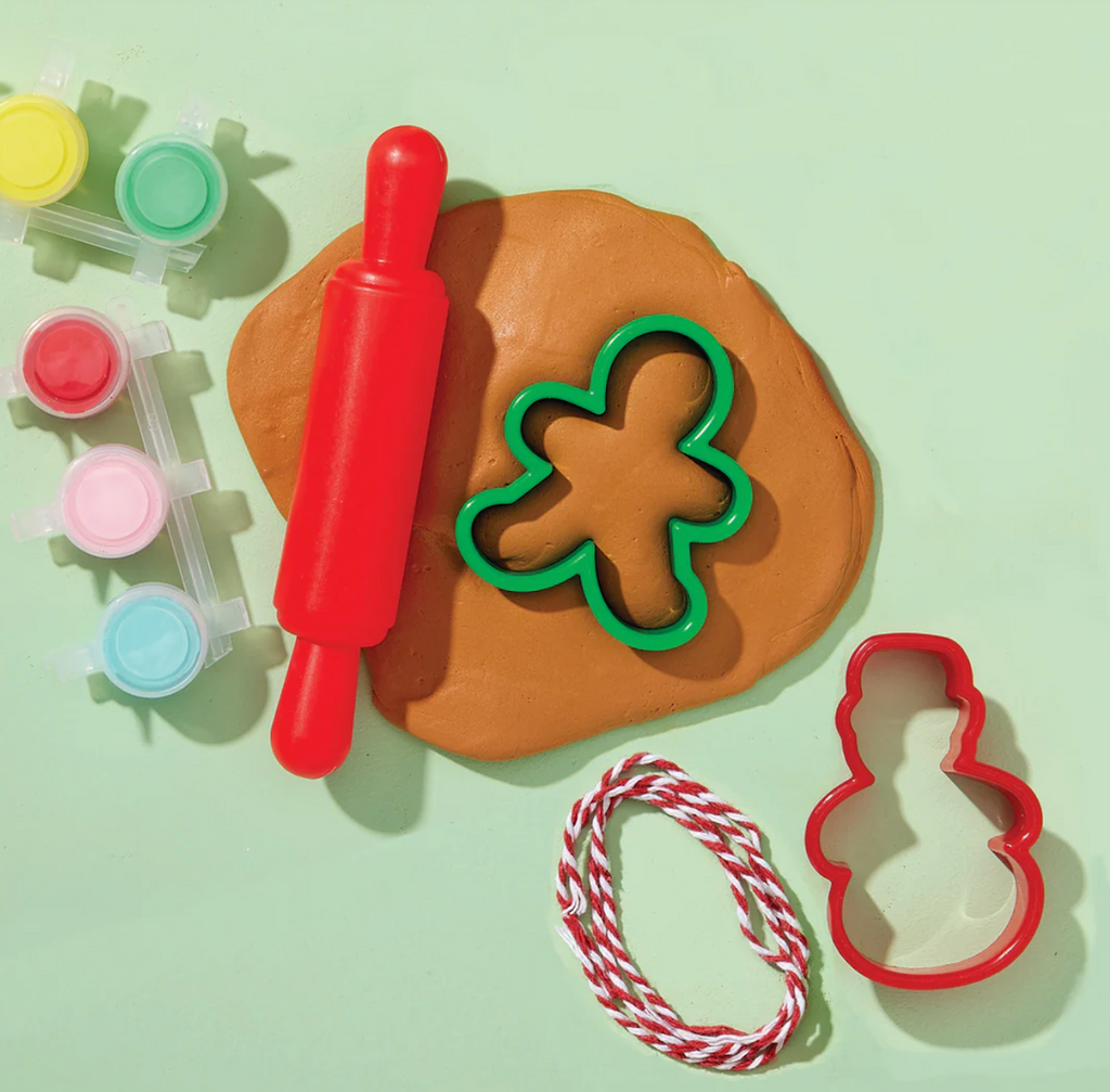 Gingerbread air dry clay rolled flat on a green surface with a red plastic rolling pin, a green gingerbread man cookie cutter in the clay, and other cutters, red and white striped string, and colorful paints. All these and more are included in ornament makign kit. 