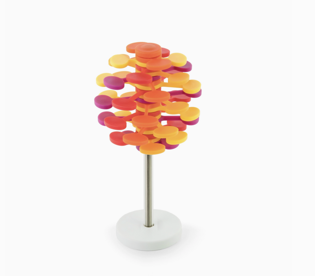 Mango Lollipopter in white stand. Lollipopter  has red, orange, yellow, and magenta plastic pieces that move when twisted on its silver base.