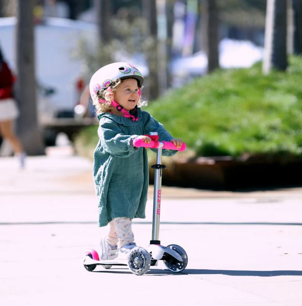Child riding the Micro Mini Flux LED Scooter in Neon Pink with a helmet on a sidewalk. 