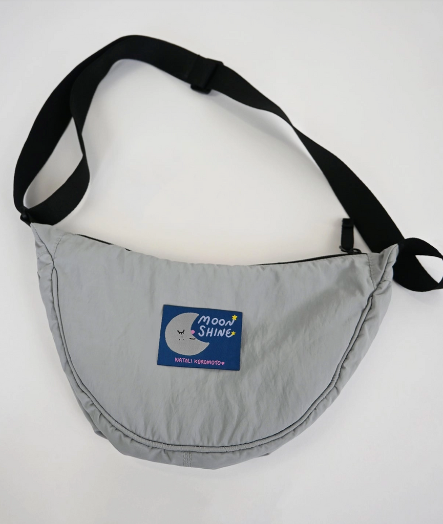 Gray crescent bag with a blue label that reads Moonshine and Natali Koromoto. 