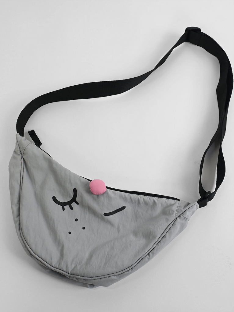 A grey crescent shaped bag with black strap that has a plush pink nose and a winking eye on the front. 