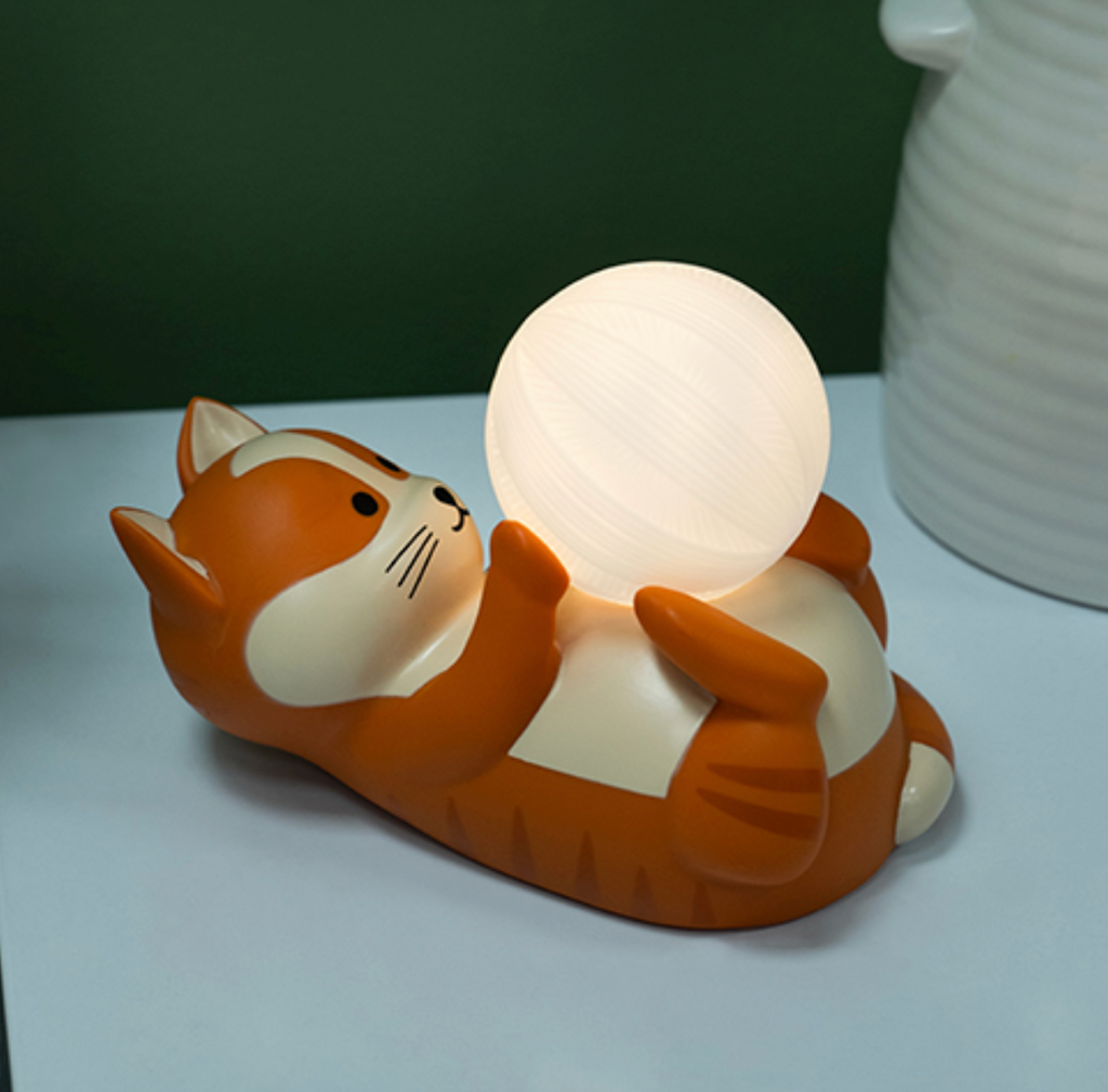 Nacho the Cat Mood Light illuminated while on a bedside table. Nacho is an orange kitty with cream colored stomach who is rolling on his back playing with a ball of yarn that serves as the light. 