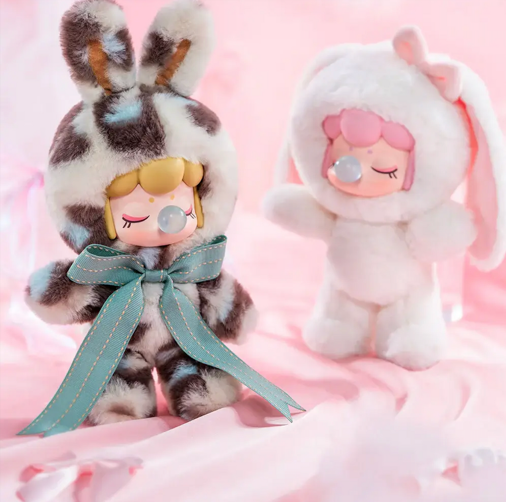 Two Nanci Shining Bunny plush figures, one with leopard print fur, bunny ears and blue bow and the other with white fur, lowered bunny ears and a pink bow on the top of her head. 