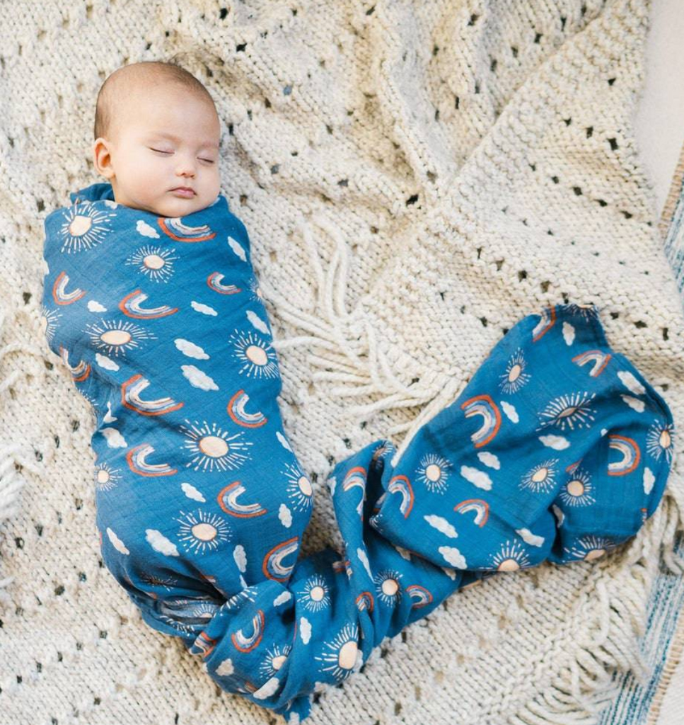 The sweetest baby swaddled securely in the Hello Sun patterned blanket with suns, rainbows and clouds on a blue background. 