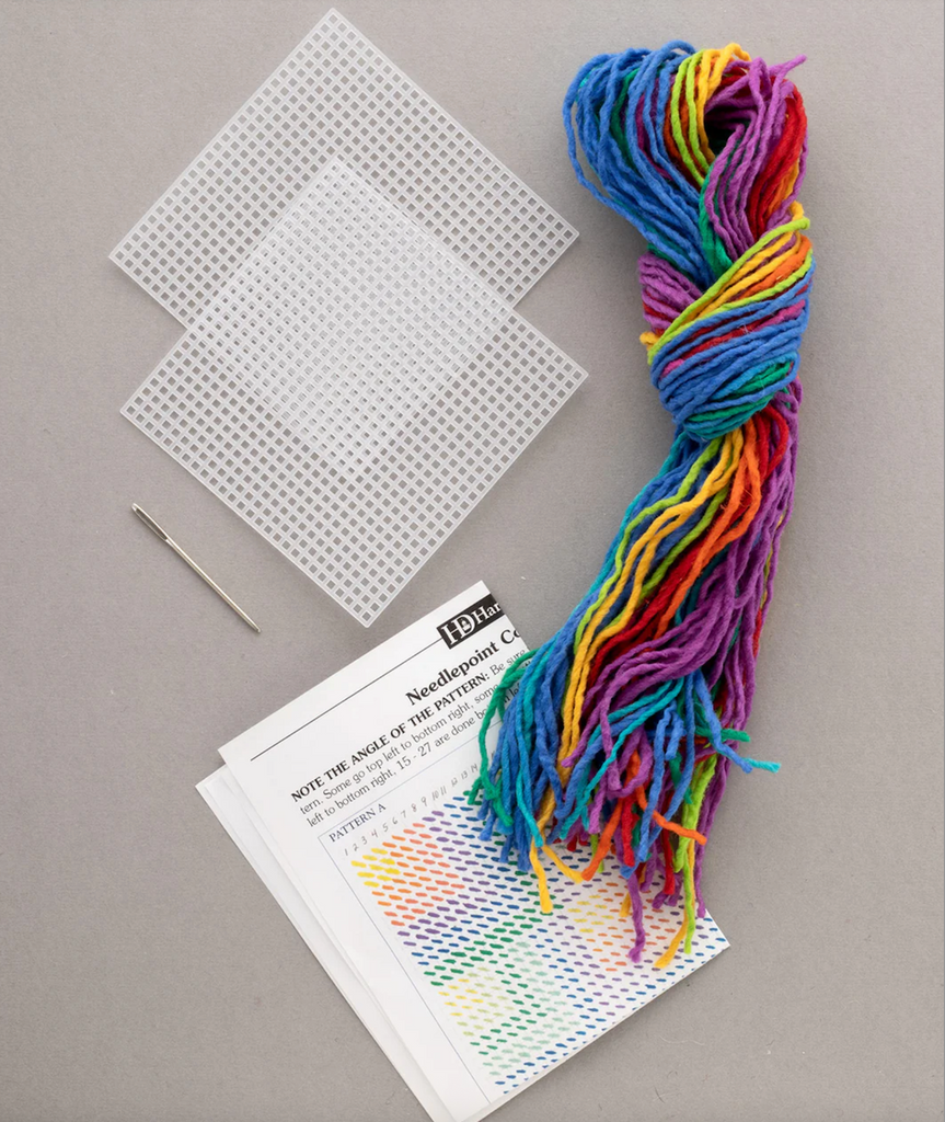 Rainbow colored yarn, needle, plastic canvases and pattern sheet that are all included in the Needlepoint Coaster Kit. 