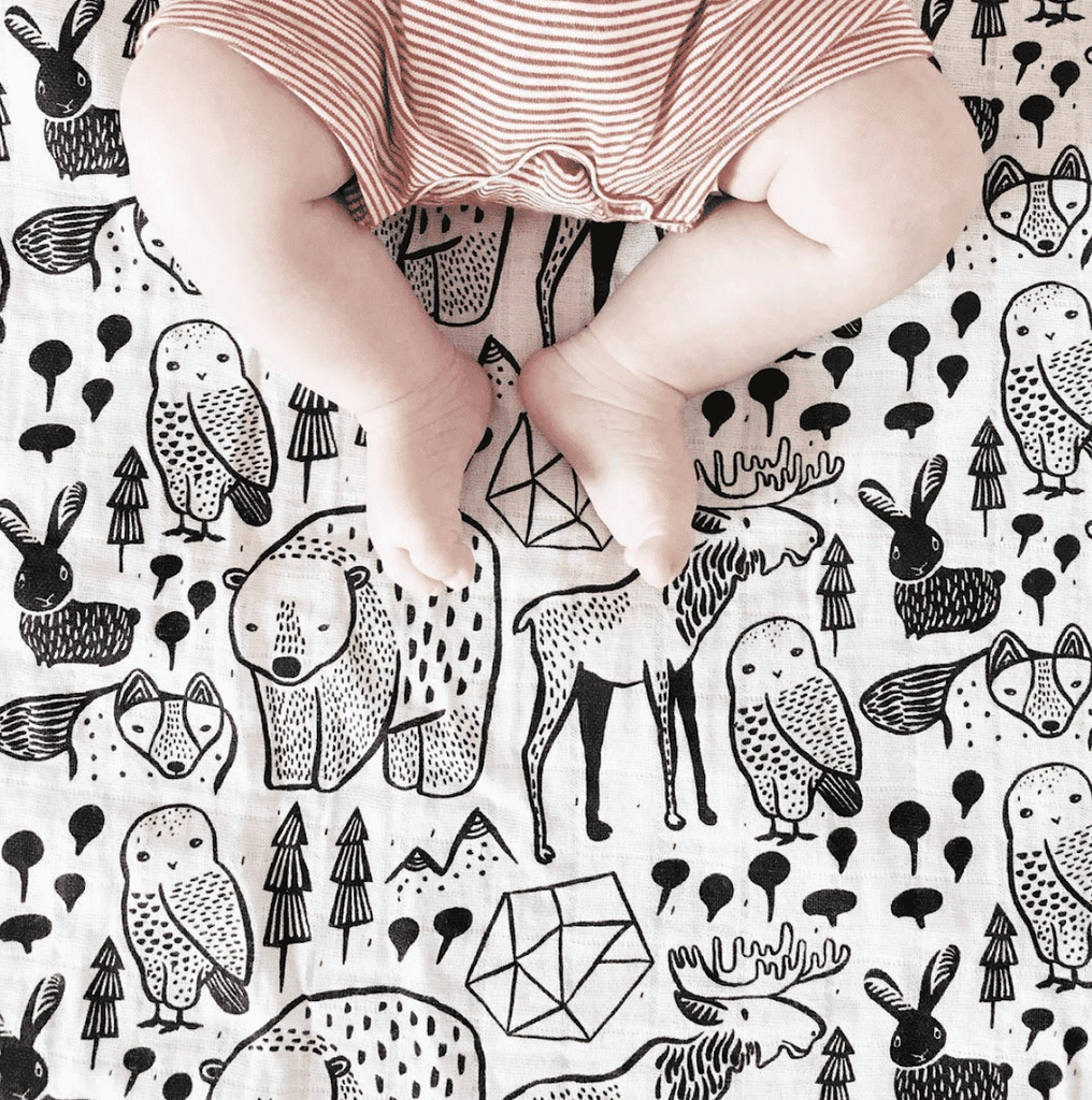 A baby's legs lying on top of the Nordic Muslin Swaddle blanket. A black and white animal pattern is printed on the blanket. 