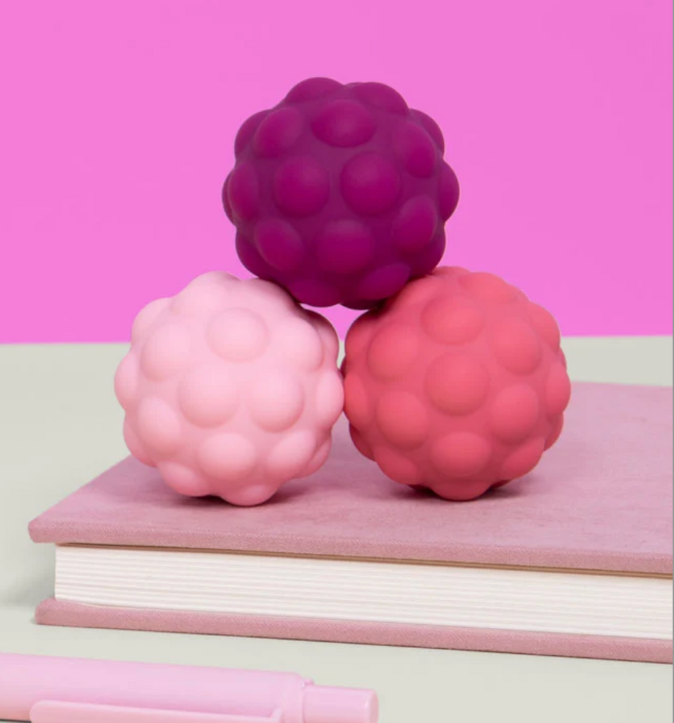 Odds Silicone Magnets. Three textured, bumpy stress balls in shades of dark magenta, light pink, and coral pink, stacked on top of a pink notebook with a pastel pink pen in the foreground. 
