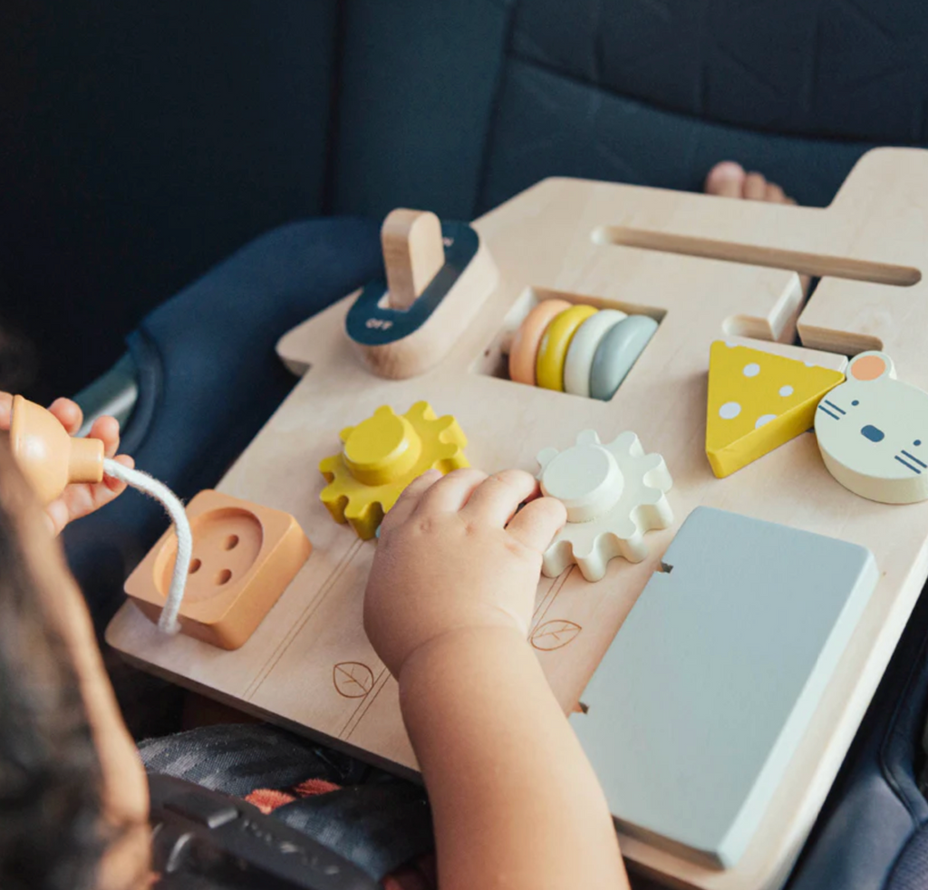 Child playing with the On the Go Busy Board wooden toy featuring various activities for toddlers to develop fine motor skills. 