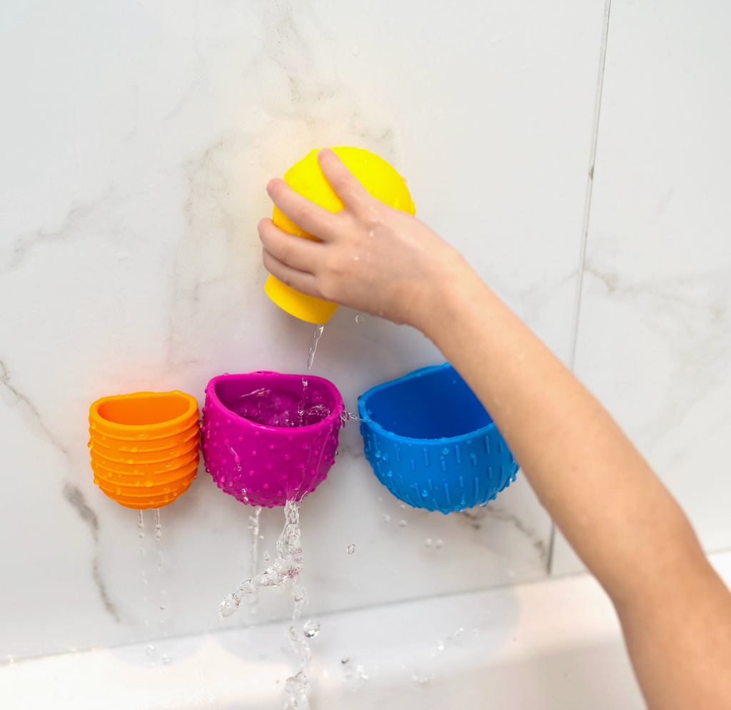 Three of the different sized brightly colored silicone pieces suctioned to the side of a bath tub with a child's hand using the silicone bulb piece to pour water into them.
