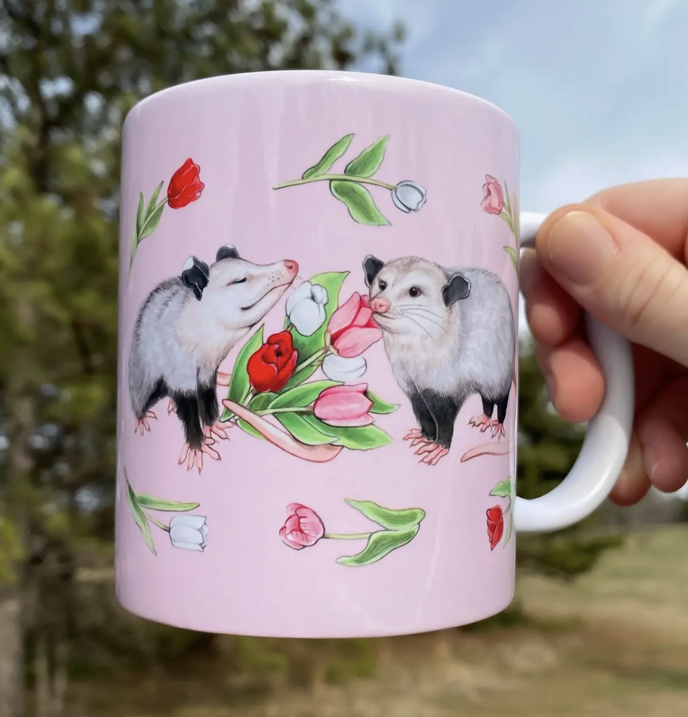 Pink mug with cute illustrations of opossums and tulips with a white handle. 