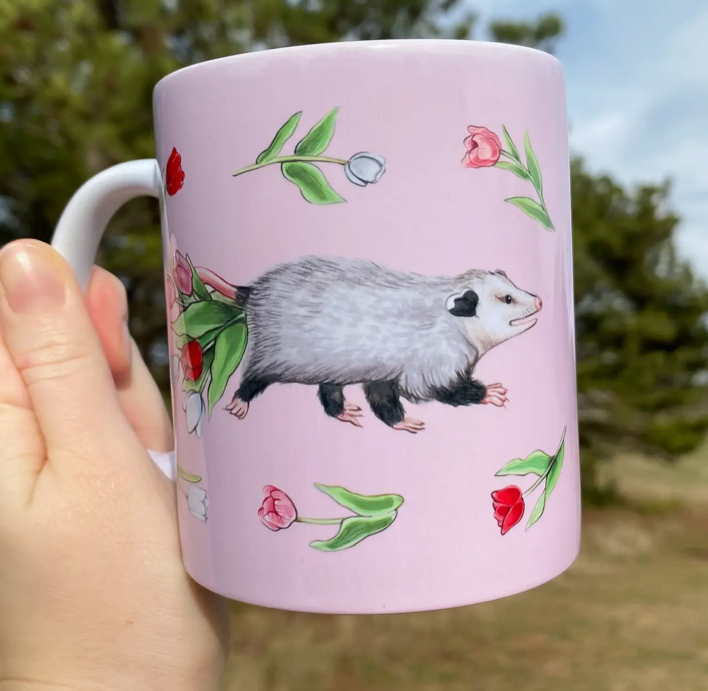 Pink mug with cute illustrations of opossums and tulips with a white handle. 