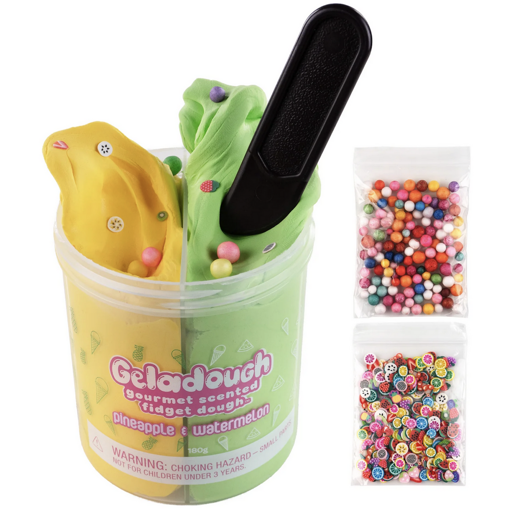 A container filled with yellow pineapple scented dough and green watermelon scented dough. With a toy spatula stuck in the watermelon scented dough. Two bags to the side of the sensory dough one filled with rainbow colored beads and the other filled with colorful confetti that looks like sliced fruits. 