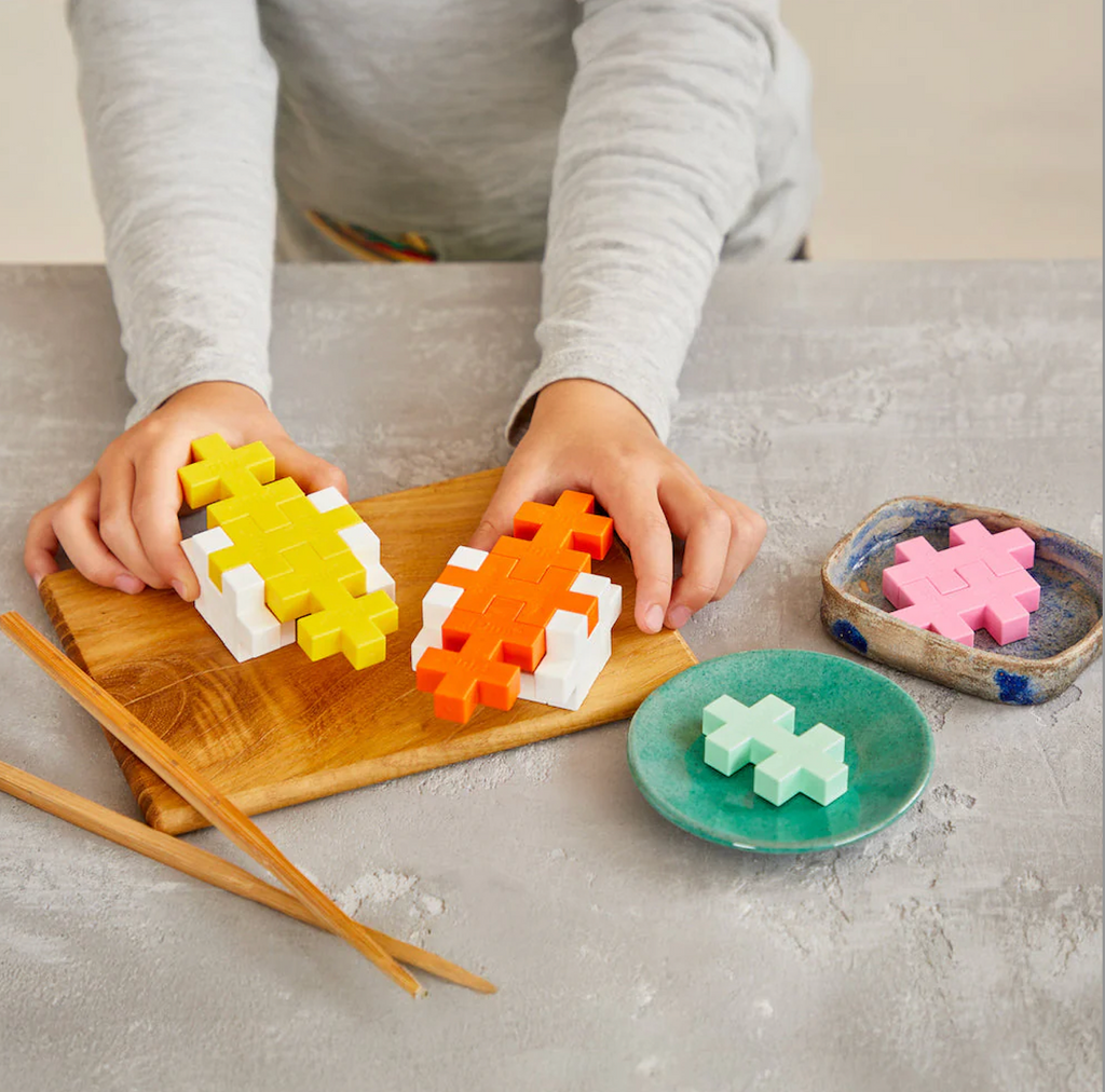 A child arranging colorful Plus Plus pieces on a wooden board built to look like sushi and other food items.
