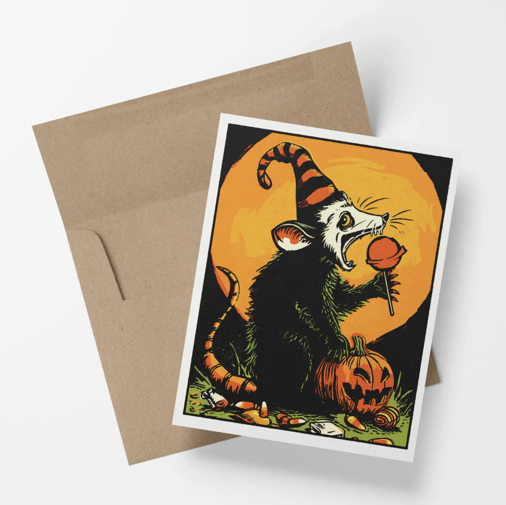 Halloween card features a possum eating a lollipop next to a jack-o-lanten in front of a glowing moon.