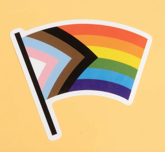 Flag shaped sticker that includes black and brown stripes to represent marginalized LGBTQ+ communities of color, along with the colors pink, light blue and white, which are used on the Transgender Pride Flag.