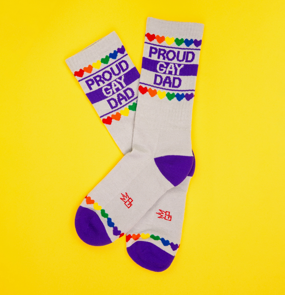 A pair of white tube socks with royal blue stripes and rainbow colored hearts that read "Proud Gay Dad" 