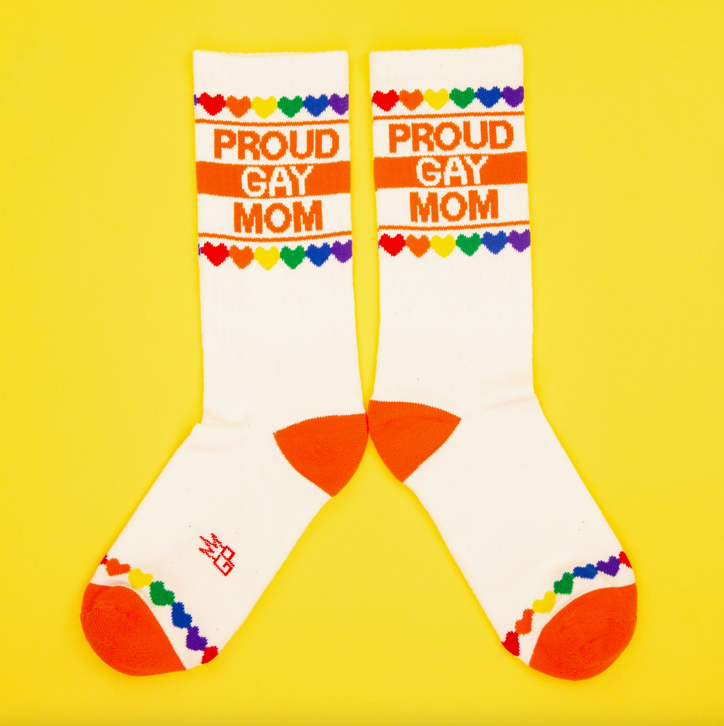 A pair of white tube socks with rainbow colored hearts and orange stripes that read "Proud Gay Mom"