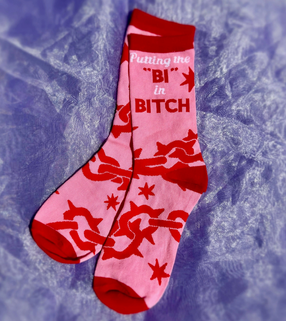 A pair of pink socks with red and white lettering that reads "Putting the 'Bi' in Bitch" with spike chain designs. 