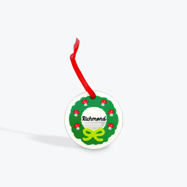 White round disc printed with a green wreath that has red mushrooms in it. Has a red ribbon for hanging. 