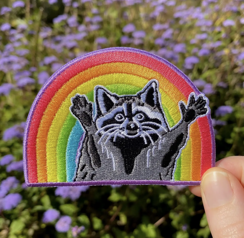 Embroidered patch with a rainbow backdrop that has a raccoon with it's arms extended in front of it. 