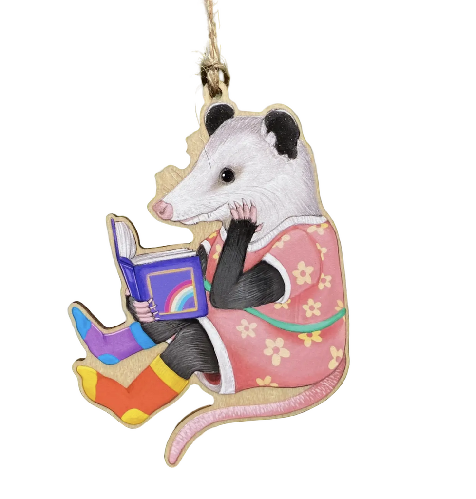Die cut wooden ornament featuring an illustration of an opossum reading a book. Wearing floral pajamas and and mismatched socks. 