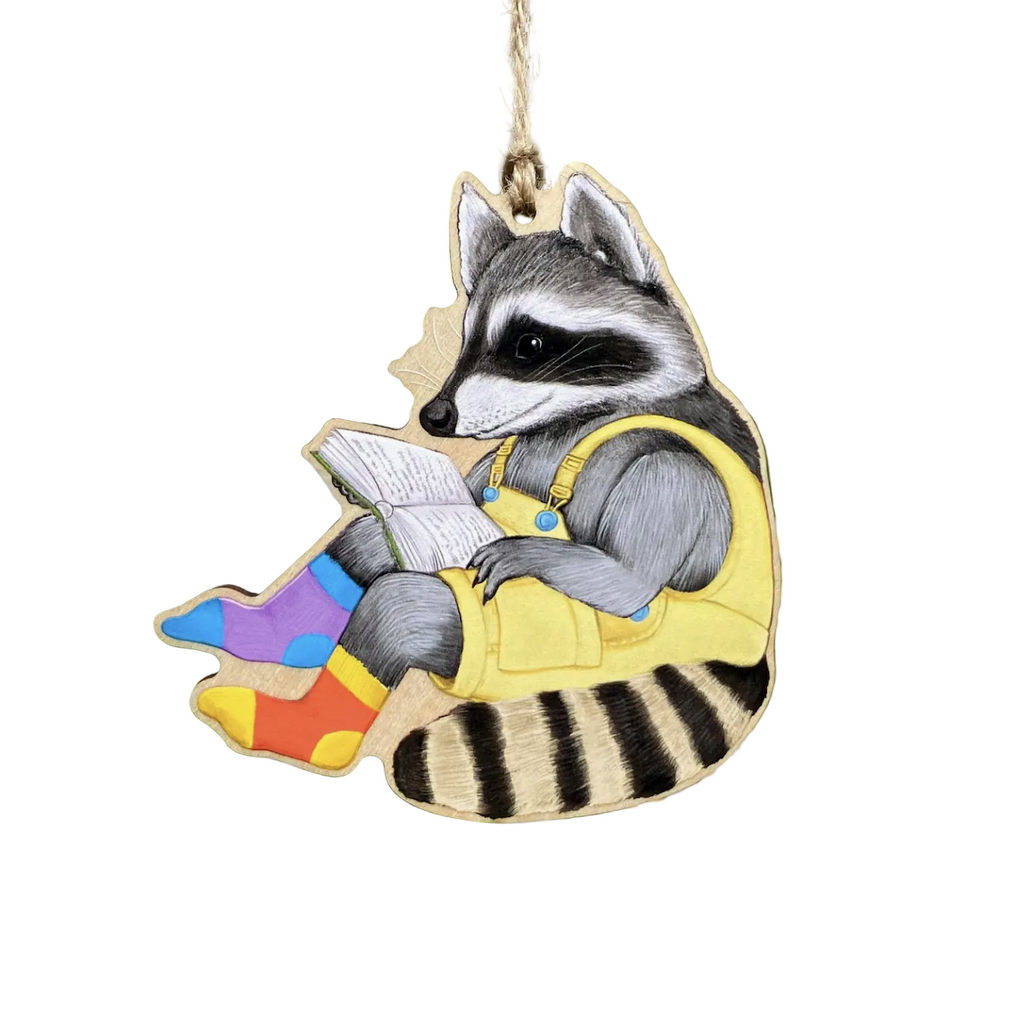 Die cut wooden ornament with a colorful illustration of a raccoon wearing yellow overalls and mismatched socks while reading a book. 