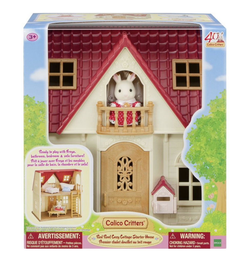 The Calico Critters Red Roof Cozy Cottage Starter Home packaged in a box with a clear window in the front showing the front of the cottage with the Freya figure perched in the top balcony. The rest of the box has pictures of the inside of the house with the included furniture. 
