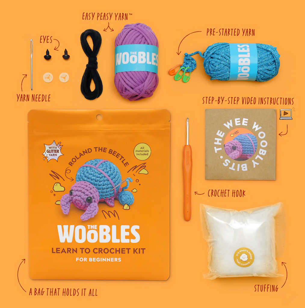 Roland the Beetle Learn to Crochet Kit with three colors of yarn, yarn needle, plastic yes, crochet hook, stuffing and instruction booklet beside the orange bag with a picture of blue and purple Roland the Beetle. 