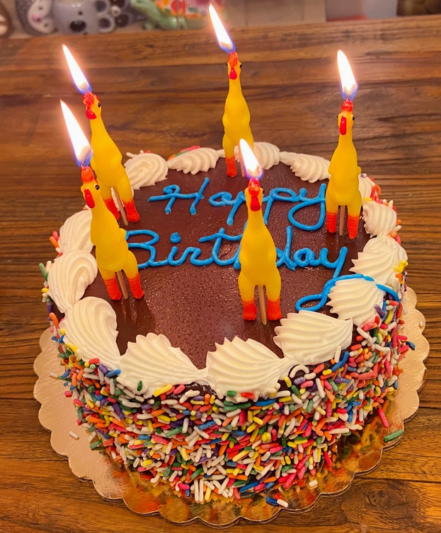 The rubber chicken candles lit on a chocolate cake that reads "Happy Birthday"
