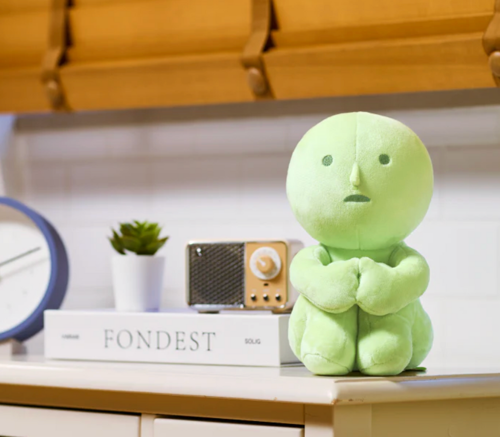 Plush green Smiski sitting on top of a desk and hugging it's knees to it's chest. 