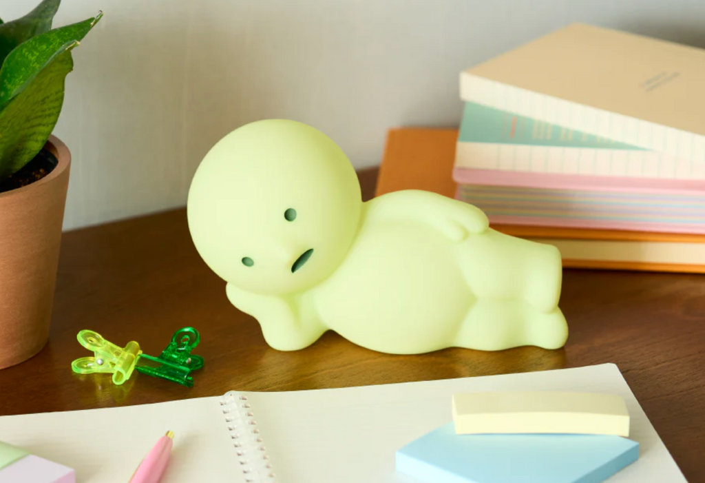 The Smiski Lounging Touch Light on top of a desk in the daylight. This shows the subtle green color of the light. 