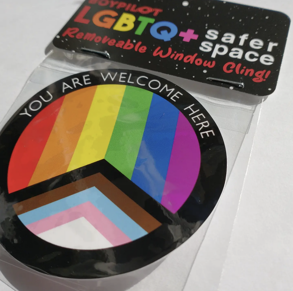 The LGBTQ+ Safer Space Window Cling packaged in clear plastic with a cardboard header. The cling of the progress flag is encircled in black and reads "You Are Welcome Here" 