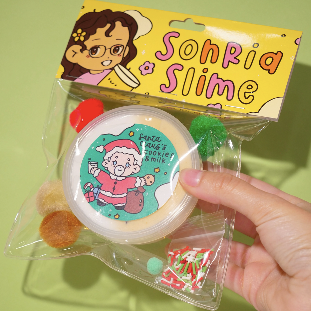 Santa Claus's Cookies and Milk slime by Sonria Slime in a clear package that includes a small tub of scented slime, red, white and green sprinkles as well as colorful pom poms. (Not food)