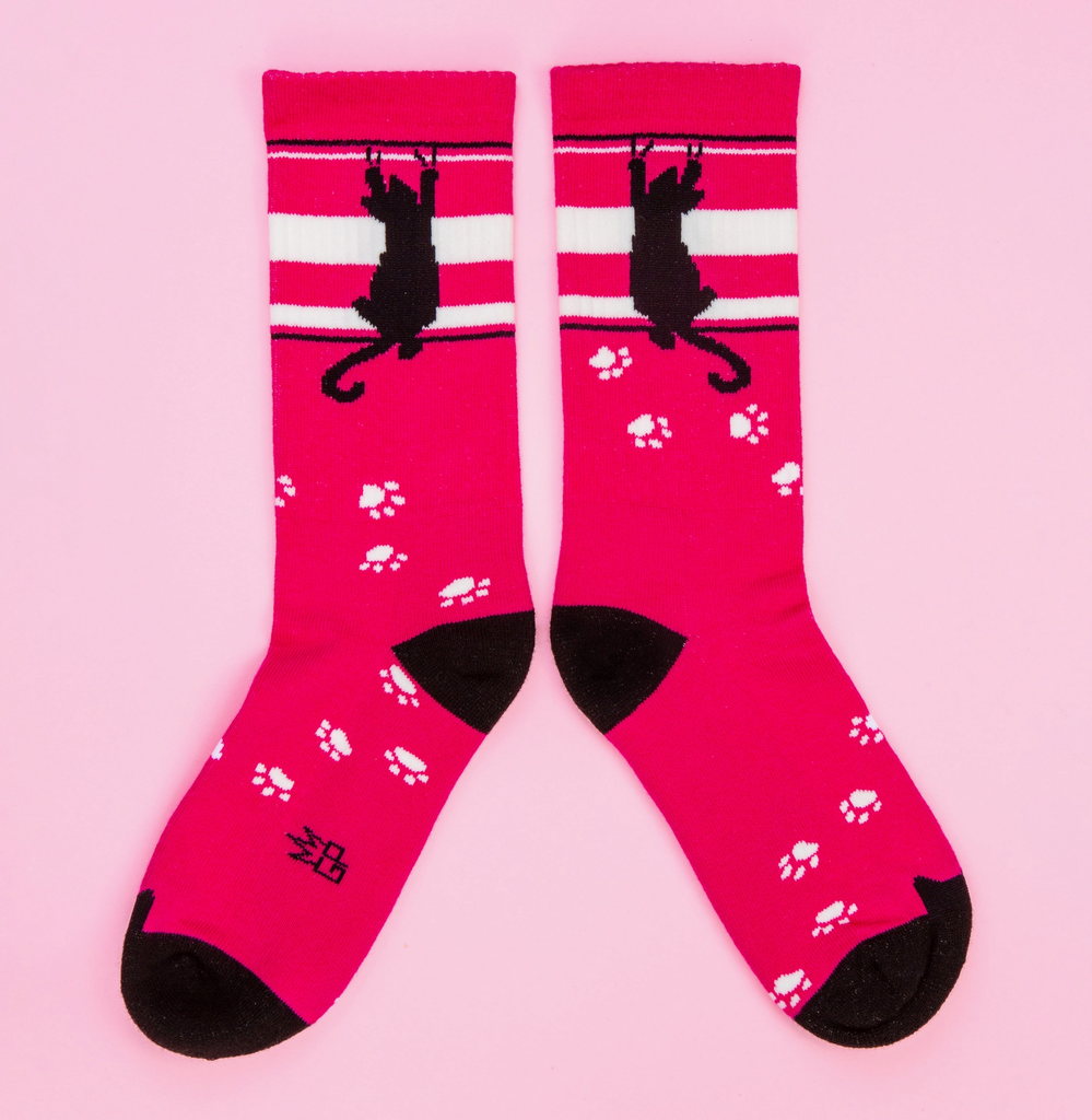 A pair of dark pink tube socks with white and black stripes, white paw prints and a black cat scratching it's way up the top of the sock. 