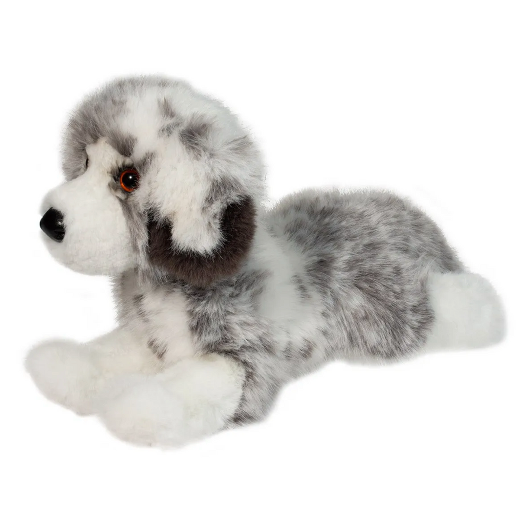 View from the left of the Australian Shepherd stuffed animal with the blue merle coat pattern. She is lying on her stomach with her white socked paws out in front.