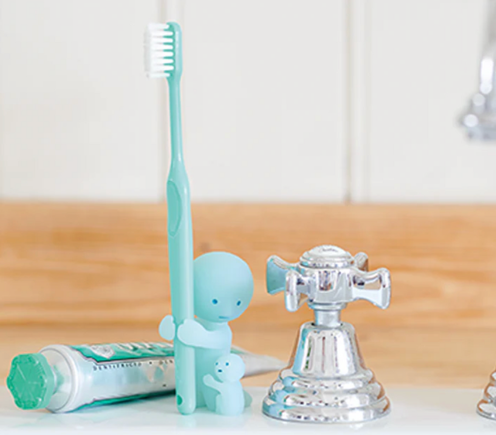 SMiski character toothbrush holder with a tiny Smiski figure hugging the leg of another figure that holds the toothbrush. They are placed on a sink beside the faucet. 