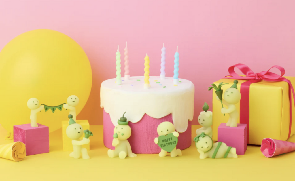 Smiski glow in the dark blind box figures from the Birthday Series celebrating 10 Years! The figures are set on a pink  and yellow background with a birthday cake, balloons and wrapped presents. 
