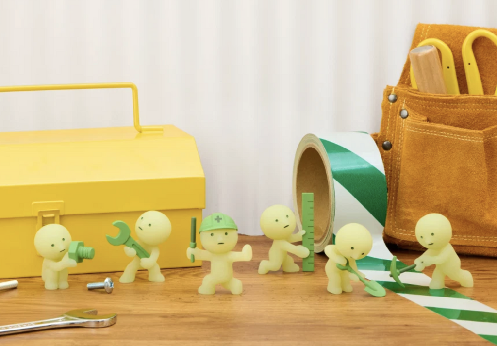 Smiski Construction series of small green construction workers holding various tools on wooden table in front of a metal toolbox.