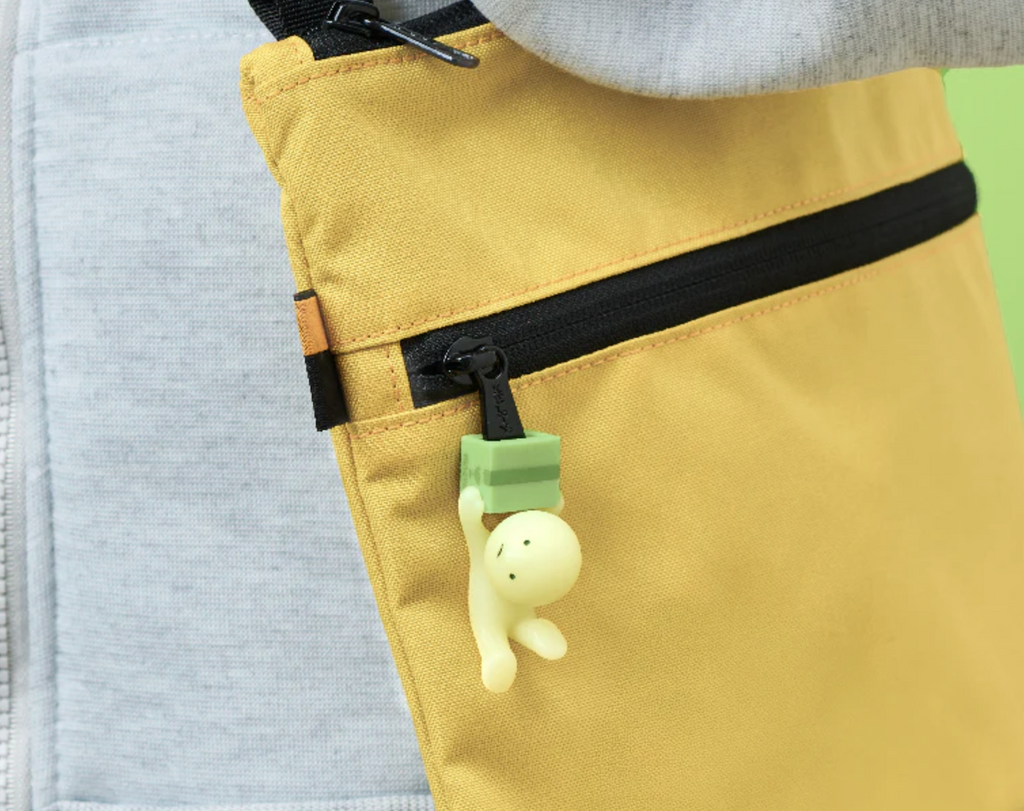 The Smiski Falling Down Zipper Bite attached to a zipper on a yellow bag. 