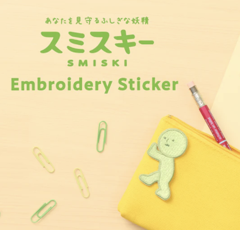 Smiski Pushing embroidered sticker on a yellow pouch near a pencil and paper clips.