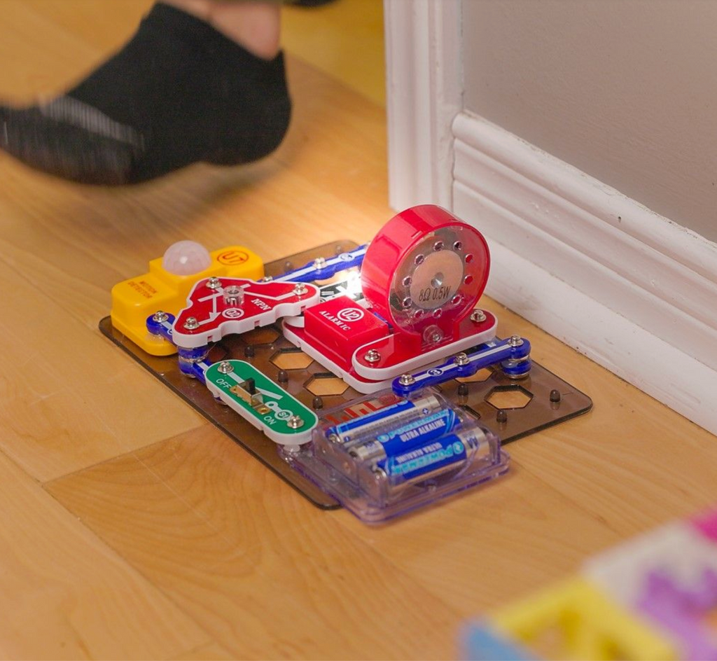 Snap Circuits circuit board built from the Spy Activity Kit placed on a wood floor, as someone walks past it setting off the trip wire. 