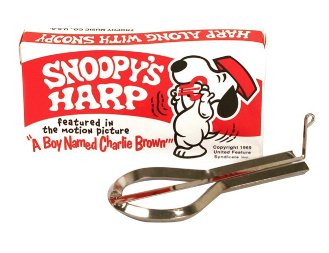A silver jaw harp musical instrument placed in front of the box it comes in which is red and white with an illustration of Snoopy with his red food bowl on his head playing the jaw harp. 