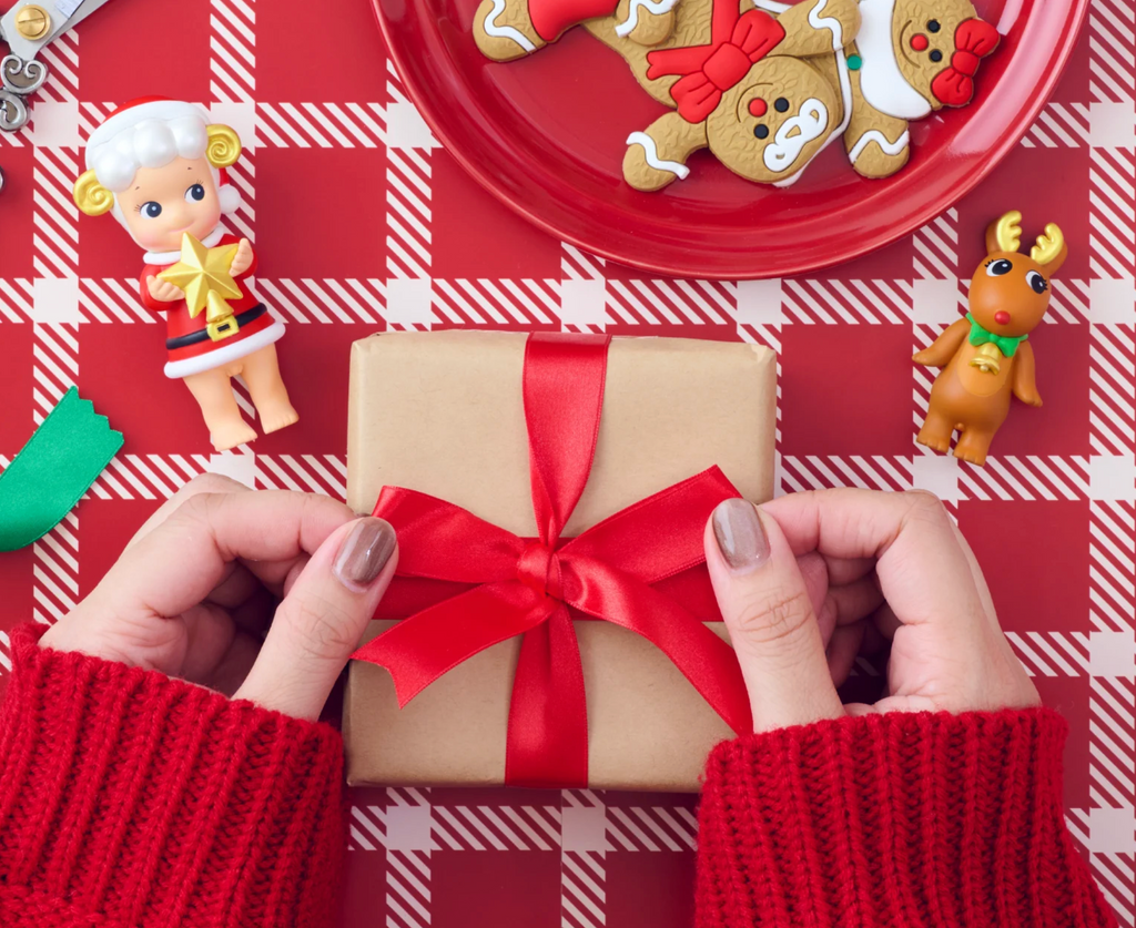 Person holding a gift box with a red ribbon on a red and white checkered tablecloth with Christmas-themed decorations with two Sonny Angels