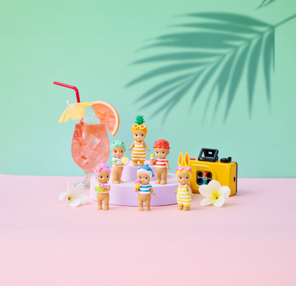A set of small toy figurines dressed in colorful striped swimwear and casual outfits, arranged on a pastel pink surface with a pink drink, yellow camera, and white flowers against a mint green background with a palm leaf shadow.