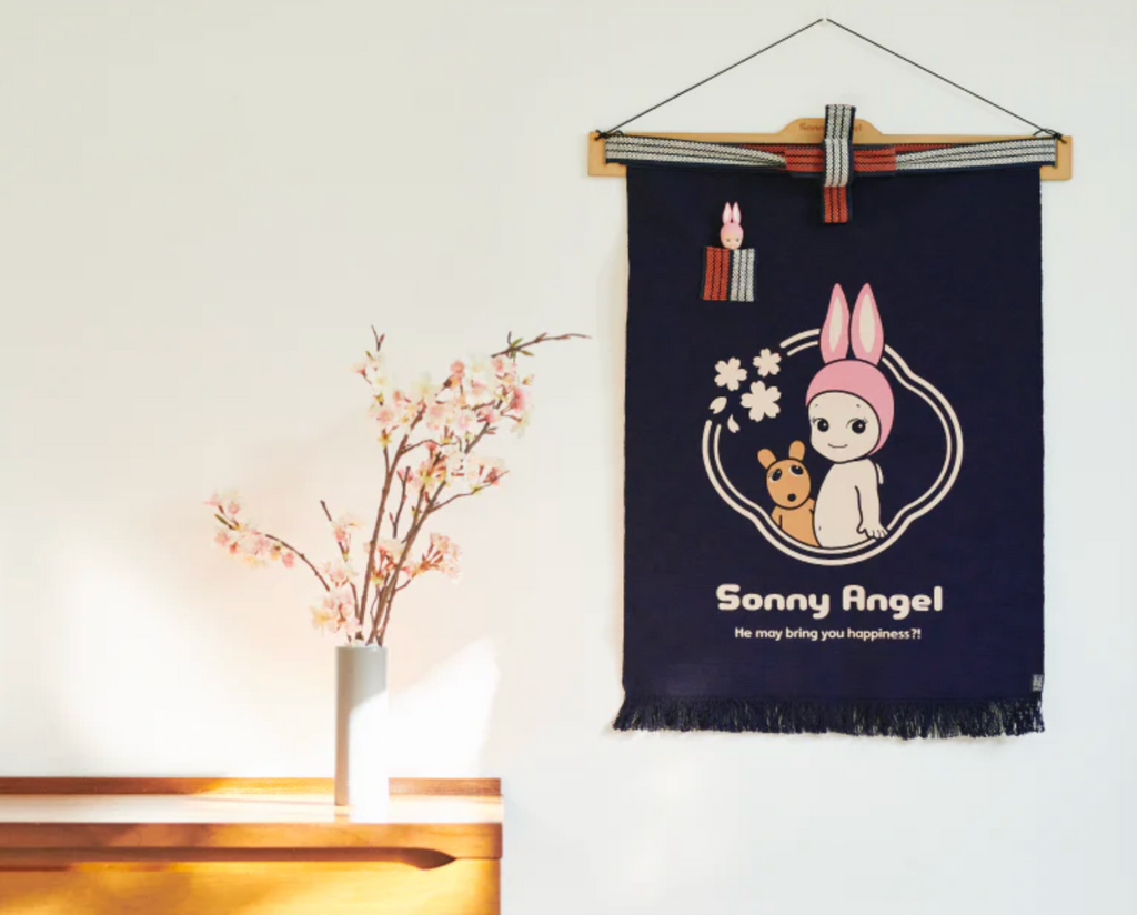 Sonny Angel maekake apron used as a wall hanging next to a wooden desk and a vase of cherry blossoms.