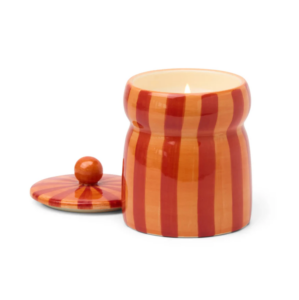 Decorative candle jar with striped pattern lit with the lid sitting to the side. 